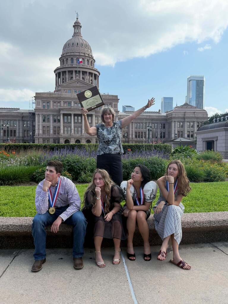 Five individuals sit on a concrete bench in front of the Texas State Capitol, posing with medals around their necks.