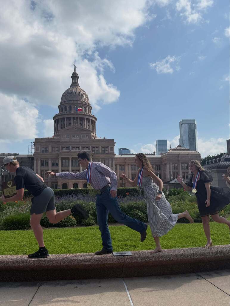 Four people run along a path in front of the Texas State Capitol building under a partly cloudy sky.