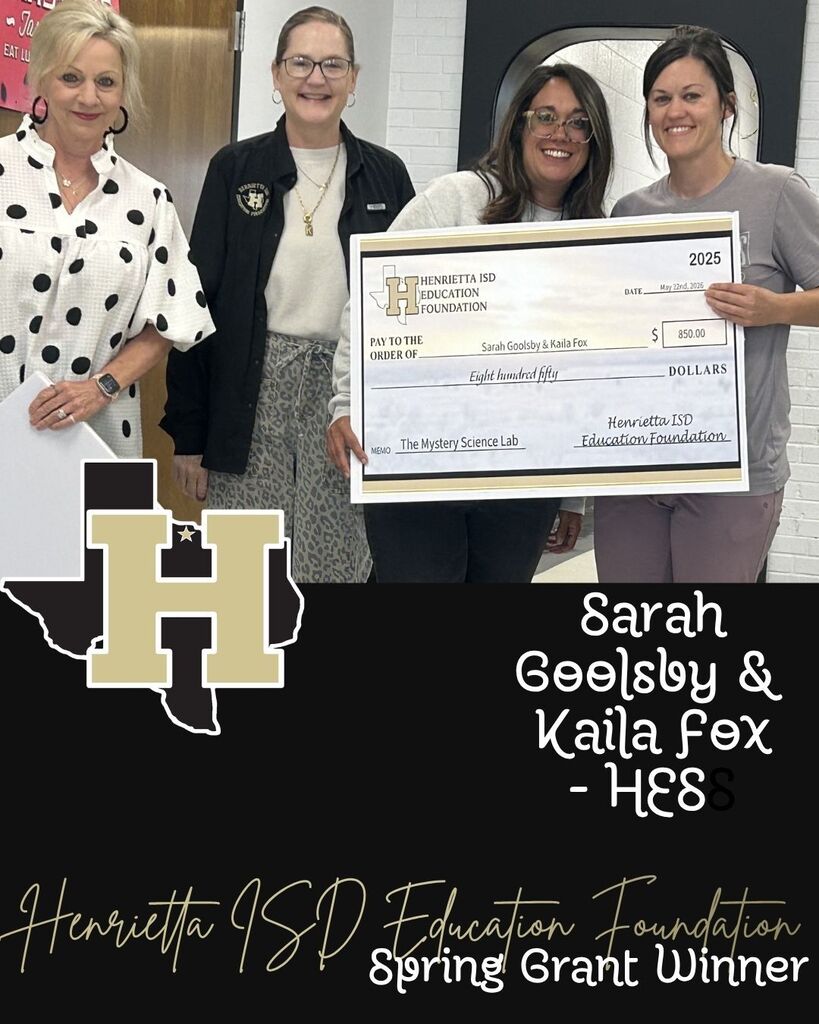 Four women pose for a picture with a large check. They stand in a hallway with a school logo.