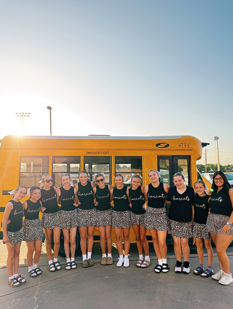 Eleven individuals in black tops and skirts stand in a line, smiling, in front of a school bus.