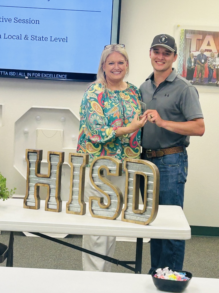 Two people stand by a table with a large sign reading "HISD". Behind them is a screen displaying a presentation.