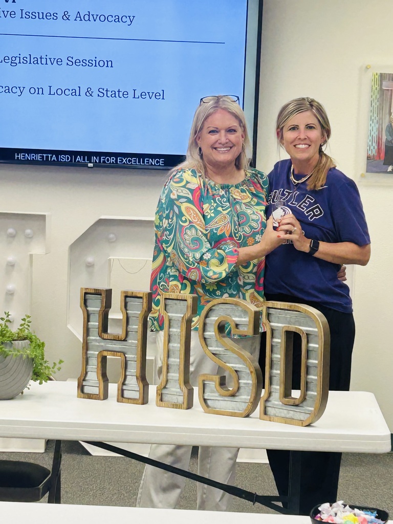Two women stand behind a desk with a sign reading "HISD" while a screen shows "Legislative Session." One holds a medal.