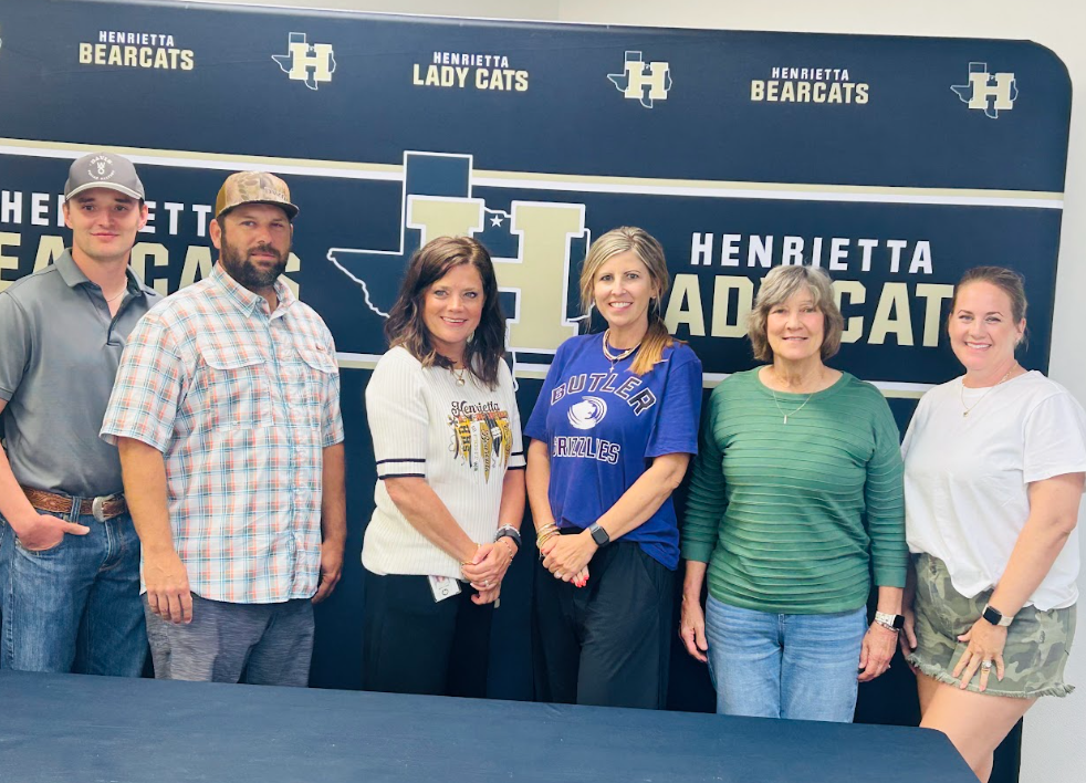Five people stand together before a wall. Three women and two men. Behind them, a banner with the words "Henrietta Lady Cats" and "H".