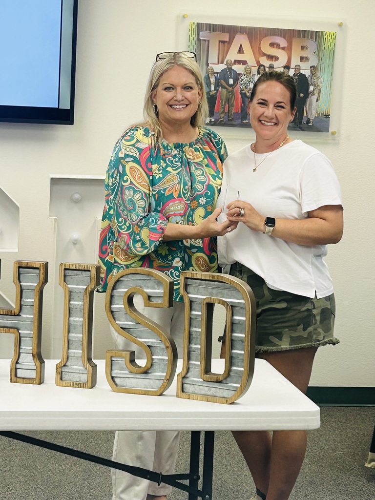 Two women stand by a table with letters "HISD" on it. One wears glasses on her head and a floral blouse.