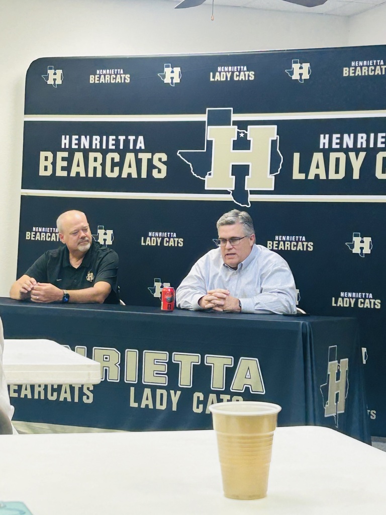 Two men sit at a table with a blue banner behind them. The banner reads "Henrietta Bearcats Lady Cats" and "Henrietta."