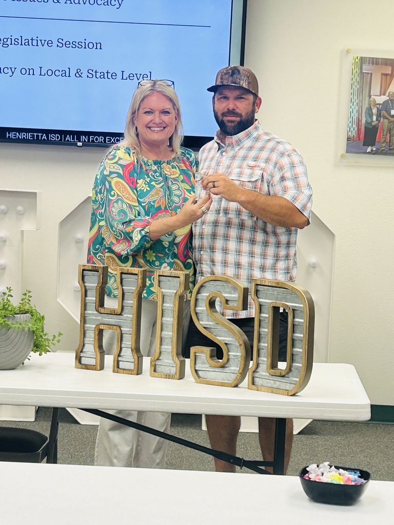 Two adults stand behind a table displaying "HISD" letters, with a plant and bowl. They smile for a photo.