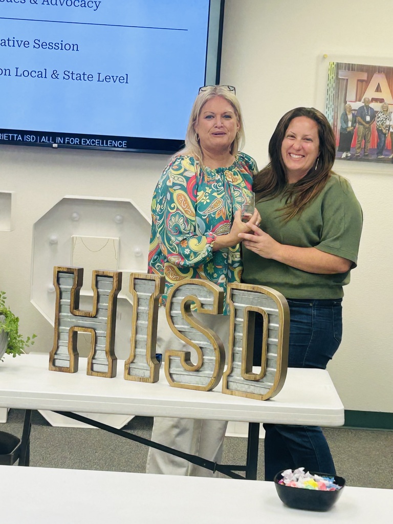 Two women stand behind a table with a sign that reads "HISD". A plant is on the table.