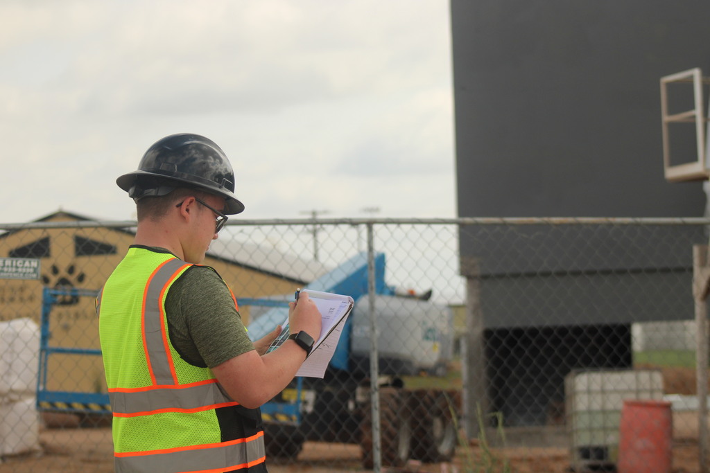 A man in a reflective vest and hard hat is writing on paper in front of a fence.