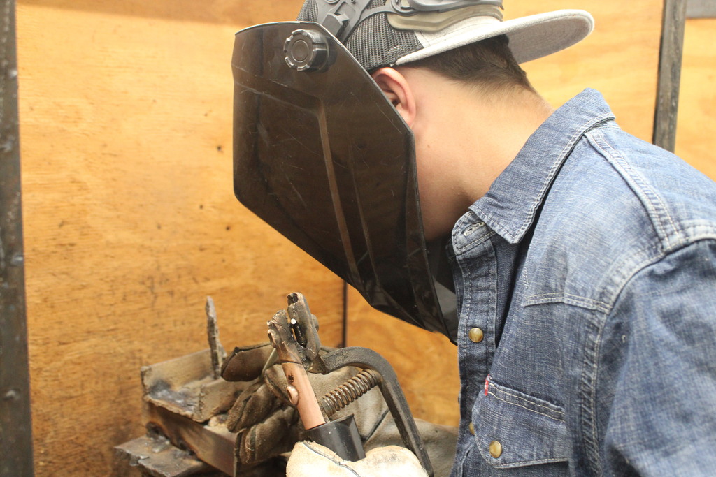 A man wearing a hat and welding mask works at a metal table. He is focused on welding.