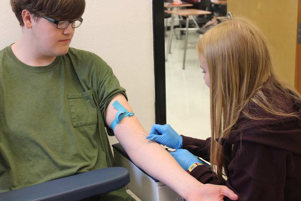 A person in a green shirt has a bandage on their arm. A person in brown assists with a blood draw.