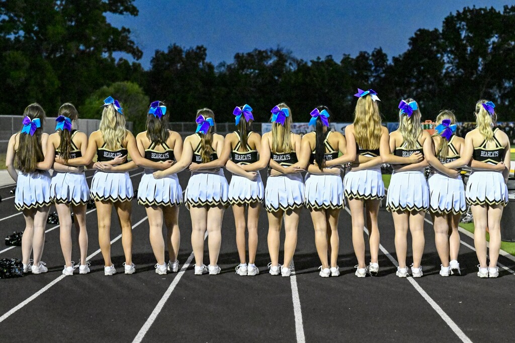 Eleven cheerleaders in matching outfits stand in a row on a track, facing away, with trees in the background.
