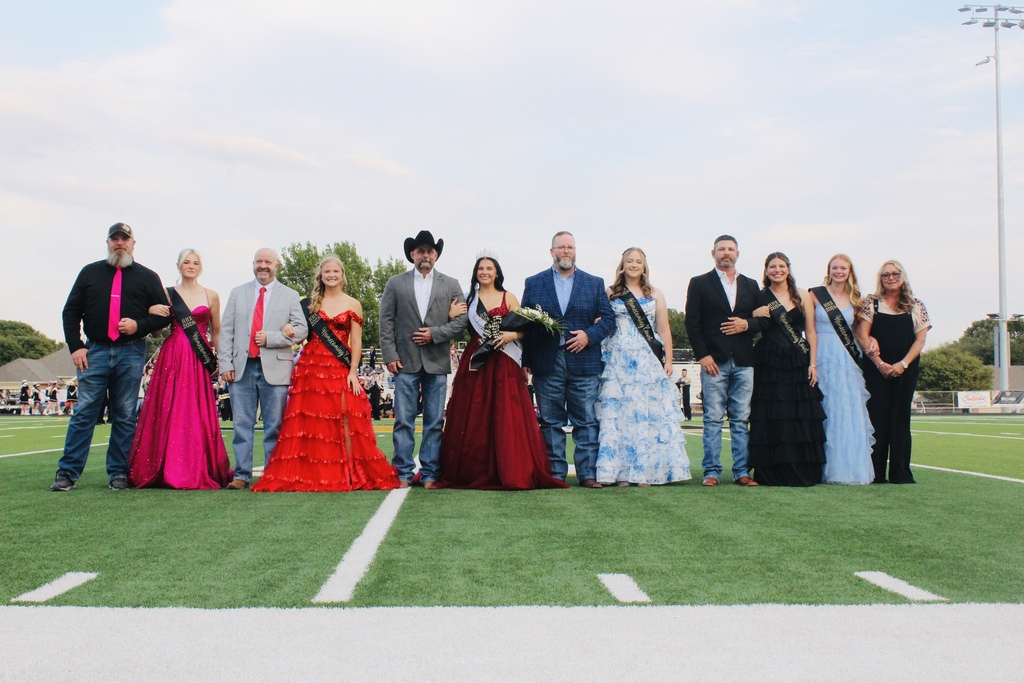 People stand on a football field. Some are in formal wear, others are in dresses. One man wears a cowboy hat.