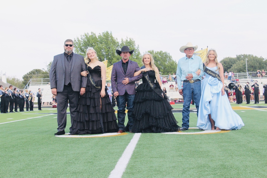 Five adults in evening wear, one in a cowboy hat, stand on a football field with a marching band in the background.
