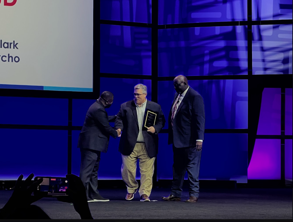 Three men, one with an award, stand on a stage; one man shakes hands with another. A screen displays text behind them.