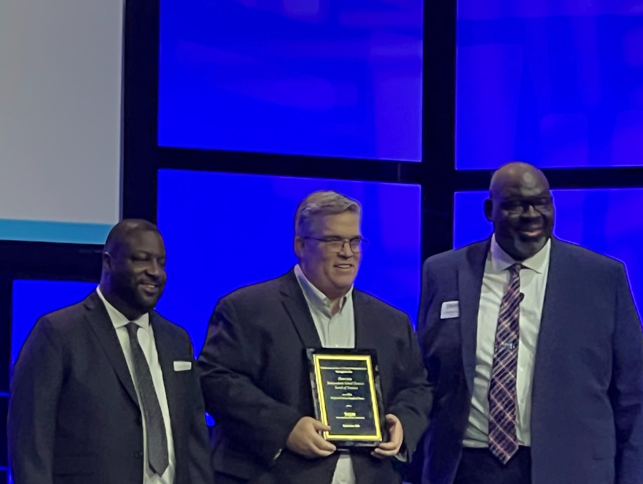 Three men in suits, one holding an award, standing together on a stage.