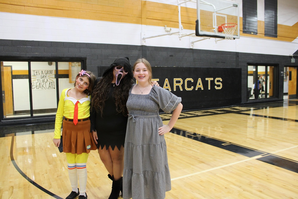 Three students in costumes pose for photo in a gym with a basketball hoop.