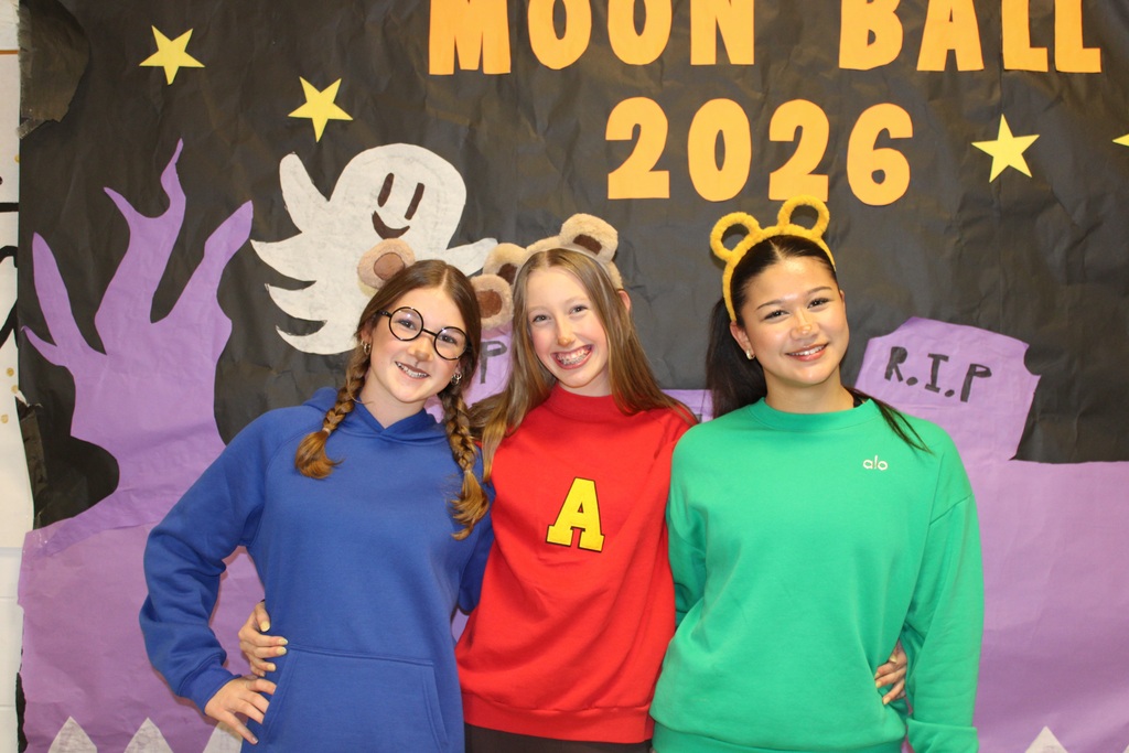 Four girls pose behind a decorated banner with a "Moon Ball 2026" title and ghostly images.