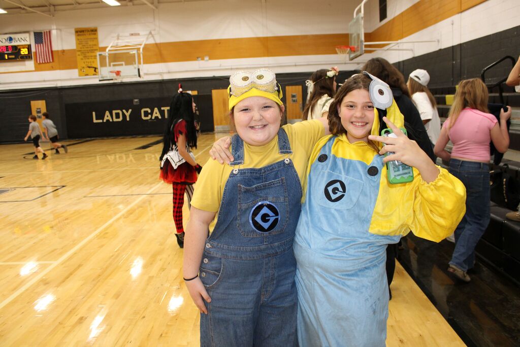 Two students dressed as minions pose in a basketball court; one holds a toy and smiles.