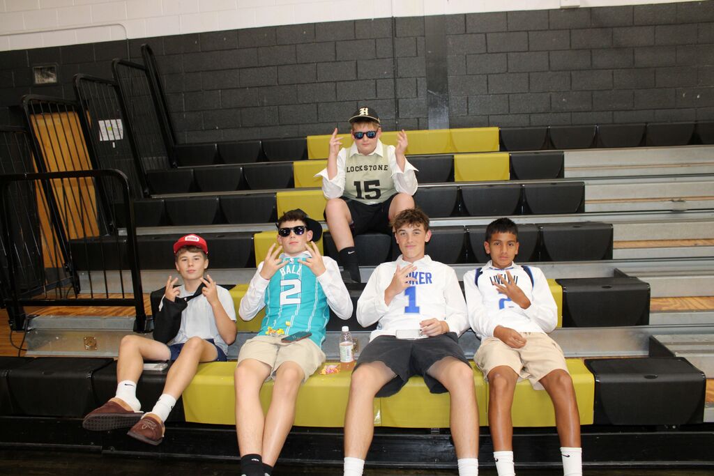 Five students seated on a yellow bleacher, posing with fingers crossed. Behind them, empty bleachers and a brick wall.
