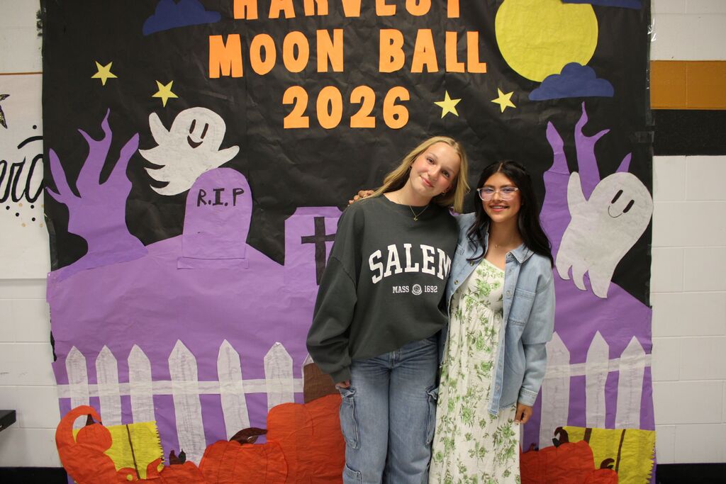 Two girls stand next to each other in front of a colorful, Halloween-themed poster.