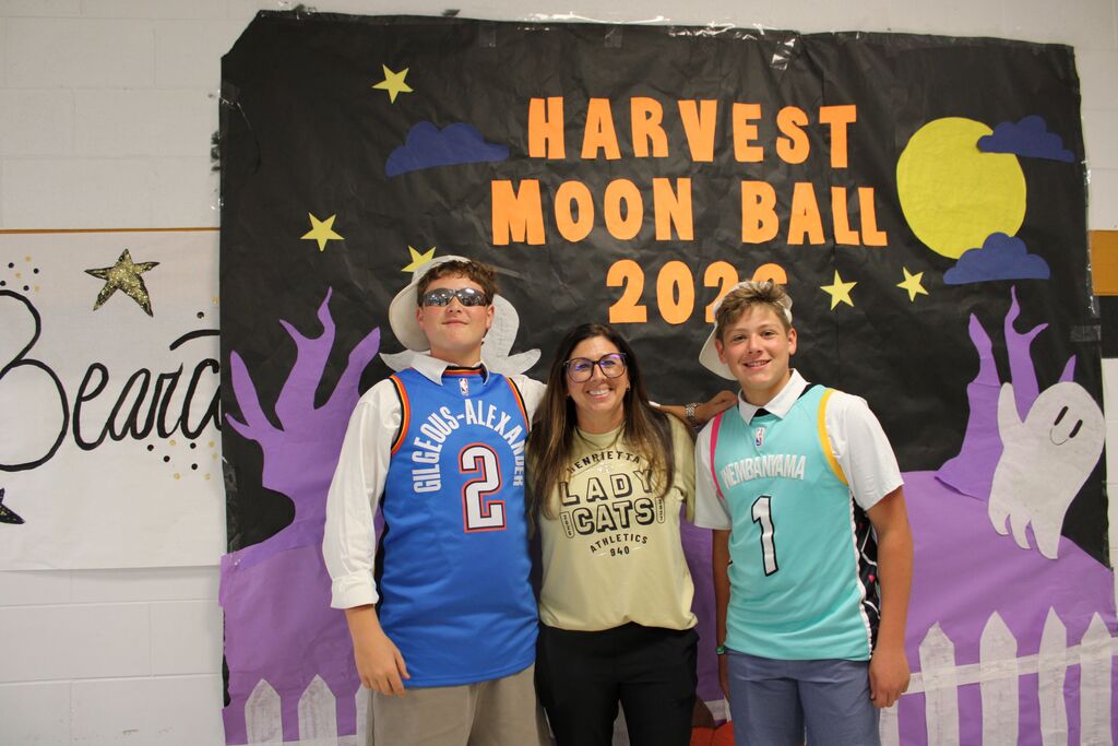 Three students stand in front of a Harvest Moon Ball 2023 banner featuring a pumpkin, moon, and stars.