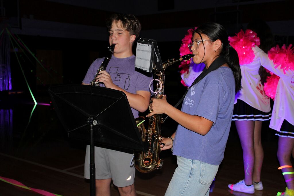 Two students play saxophones on a stage, one with a music stand. Behind them, three cheerleaders stand in pink and white uniforms.