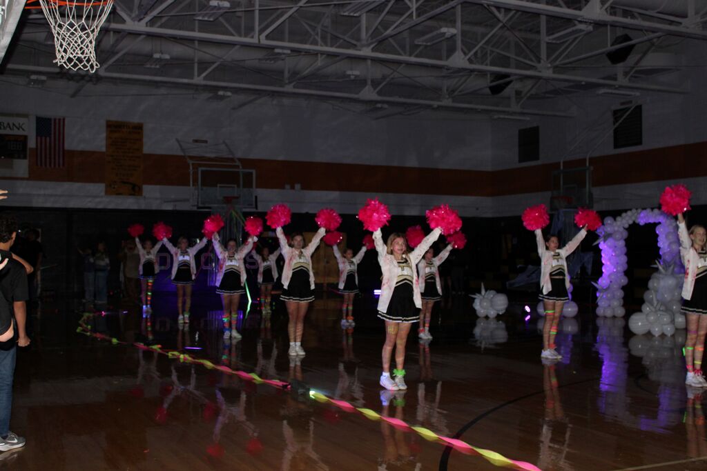 Group of cheerleaders holding pink pom-poms on a basketball court with lights and balloons.