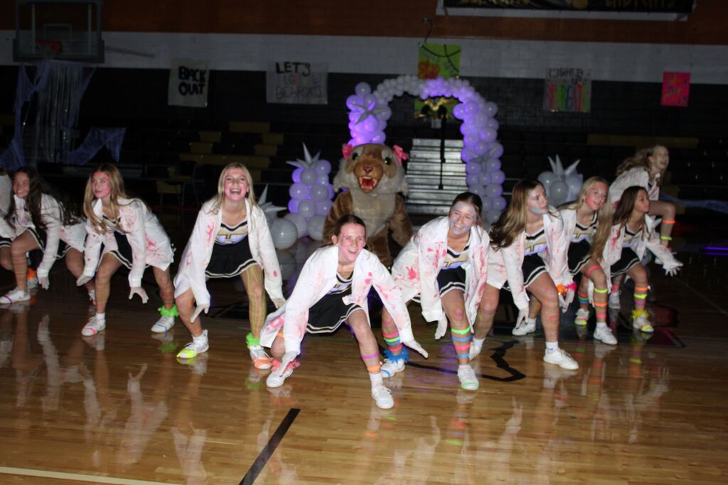 Students in white shirts and shorts, some with socks and shoes, performing dance moves on a basketball court.