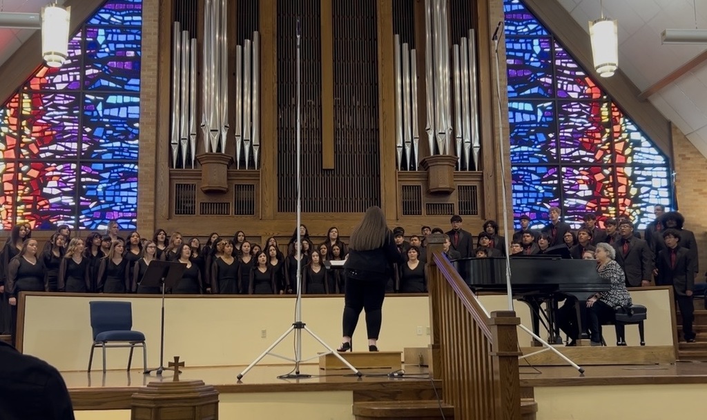 A choir in black stands before a piano. The stage has a microphone stand. Behind them are stained glass windows.