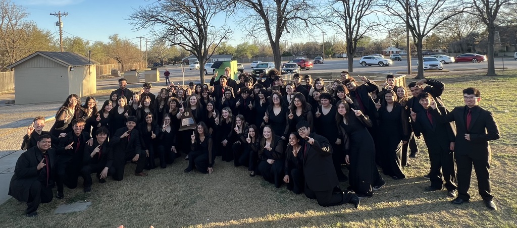 A large group of people in black attire pose for a photo on grassy land. Trees and parked cars are in the background.