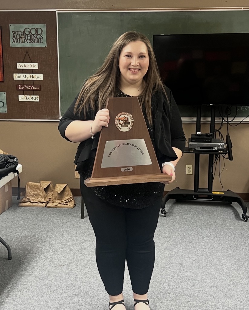 A woman poses for a photo while holding a wooden plaque in a classroom. Behind her, a TV is on a stand.