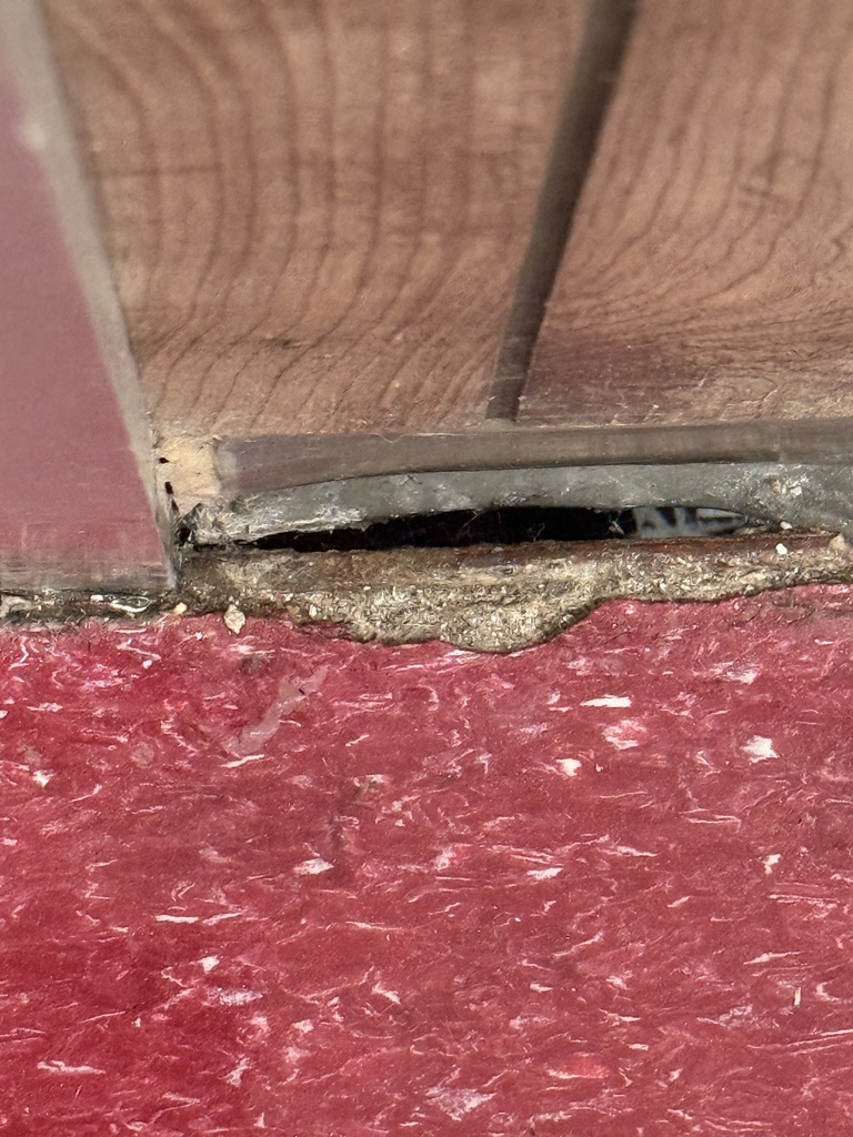 A close-up view of a wooden floor, partially covered by a red floor, with visible gaps and a damaged edge.