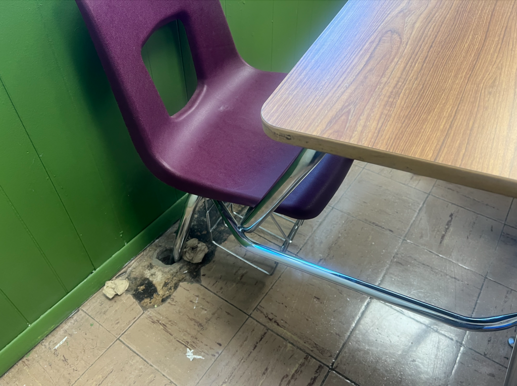 A purple chair with a hole in the back is beside a wooden desk. The chair is partly beneath the desk. The floor has tiles.