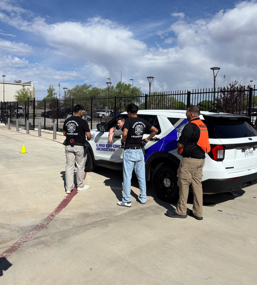 Four people stand near a white SUV with blue trim. Two wear backpacks. One has a yellow cone nearby.