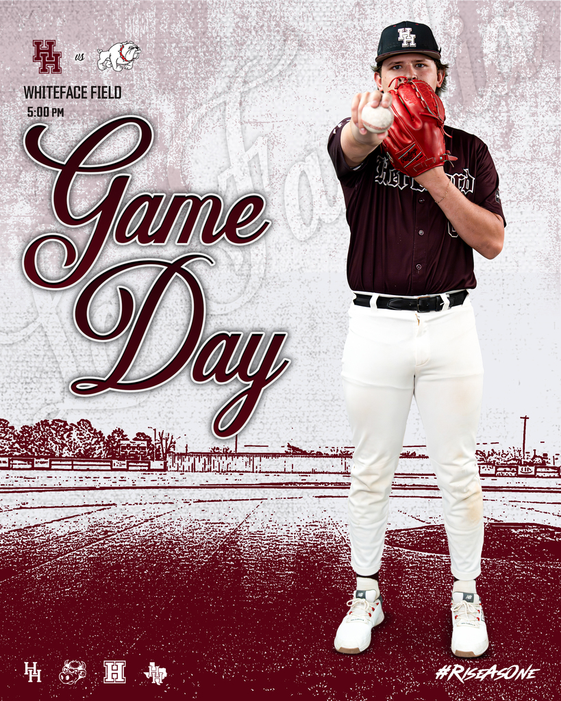 A baseball player in a maroon shirt and white pants stands on a field with a baseball.