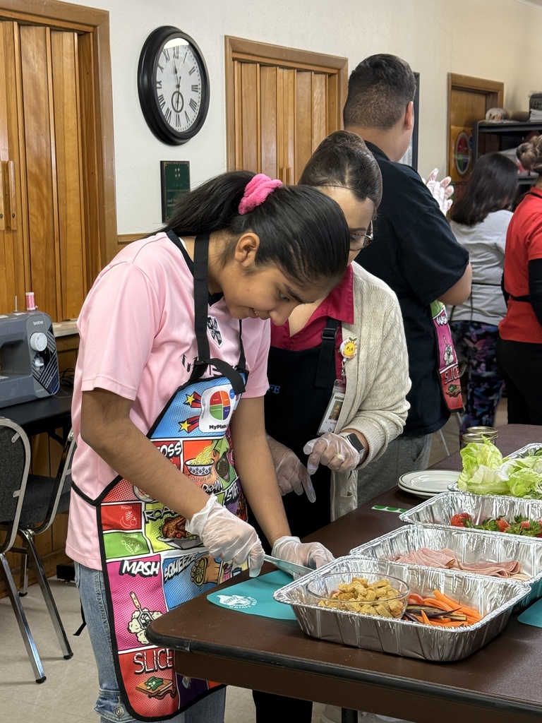 Two people, one in a pink shirt, prepare food at a table. A clock is on the wall.
