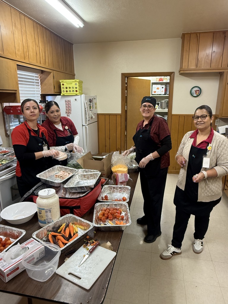 Four people in aprons stand behind a kitchen table with various foods in containers. Behind them, cabinets and a refrigerator are visible.