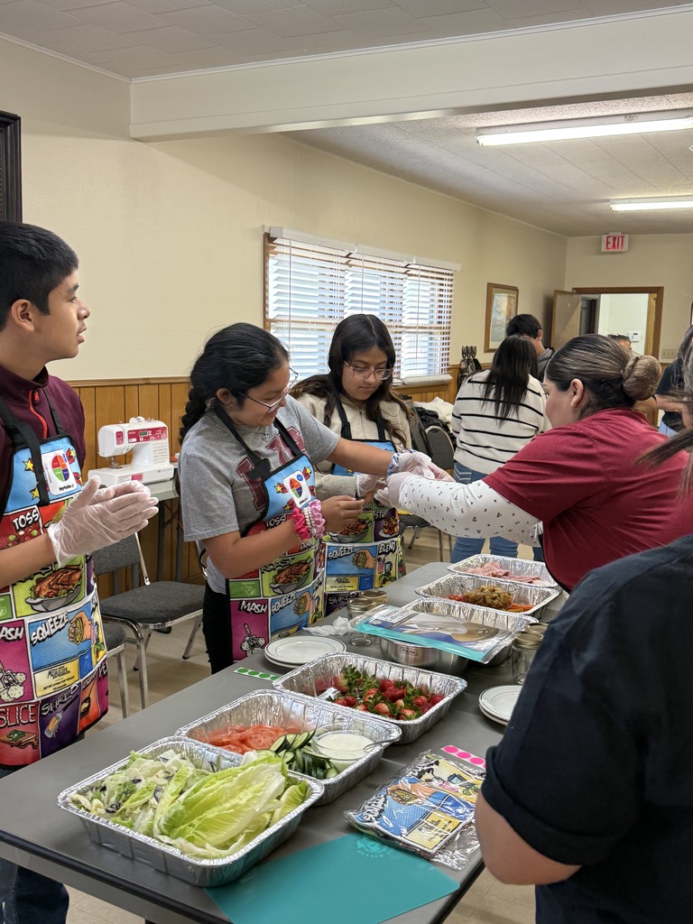 People wearing aprons prepare food in a room with windows. A table has various foods in containers.