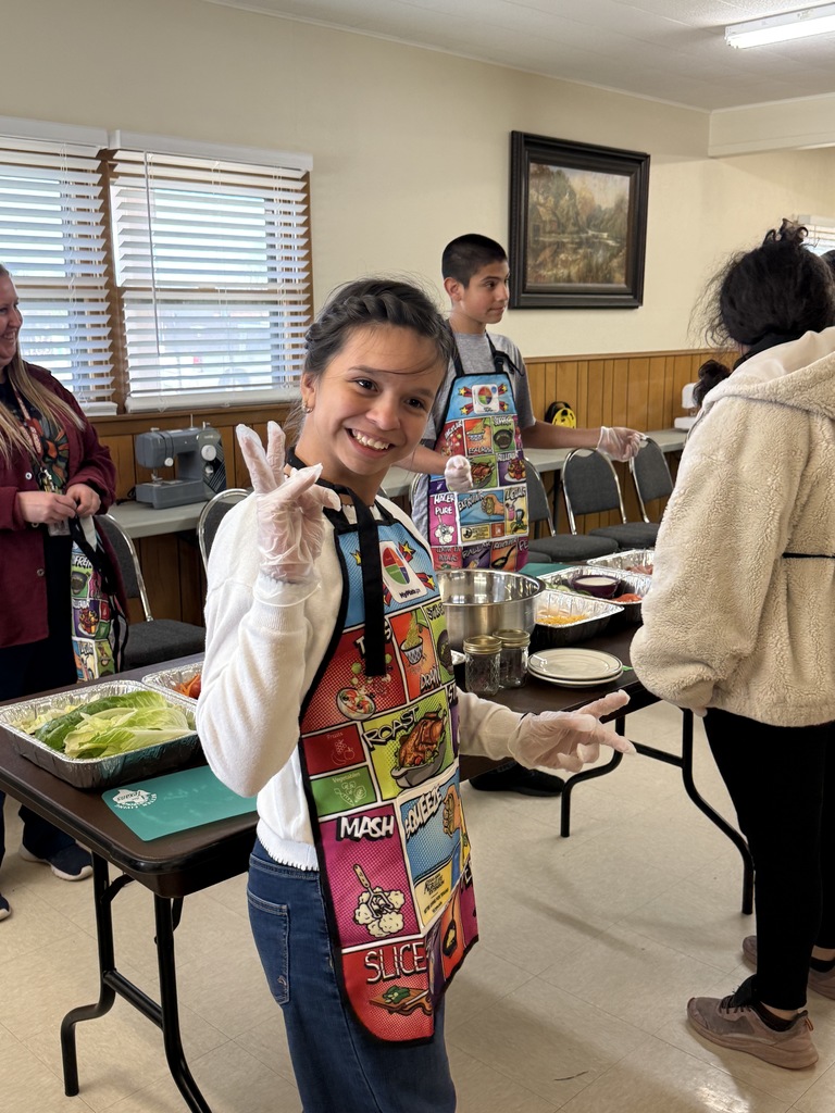 Woman in apron smiling, standing next to table with food, surrounded by other people.