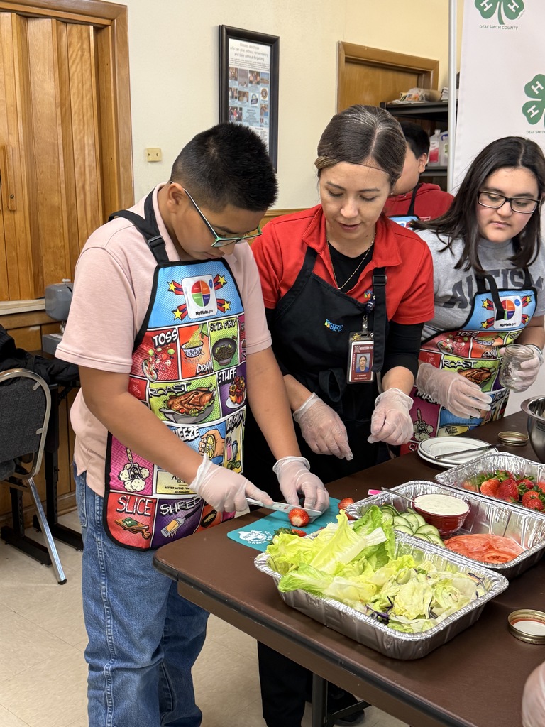 Three people are preparing salad in a classroom. A boy is standing with a colorful apron.