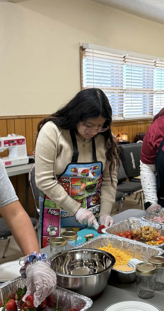 Two people wear aprons and gloves to prepare food at a table with bowls, plates, and jars.