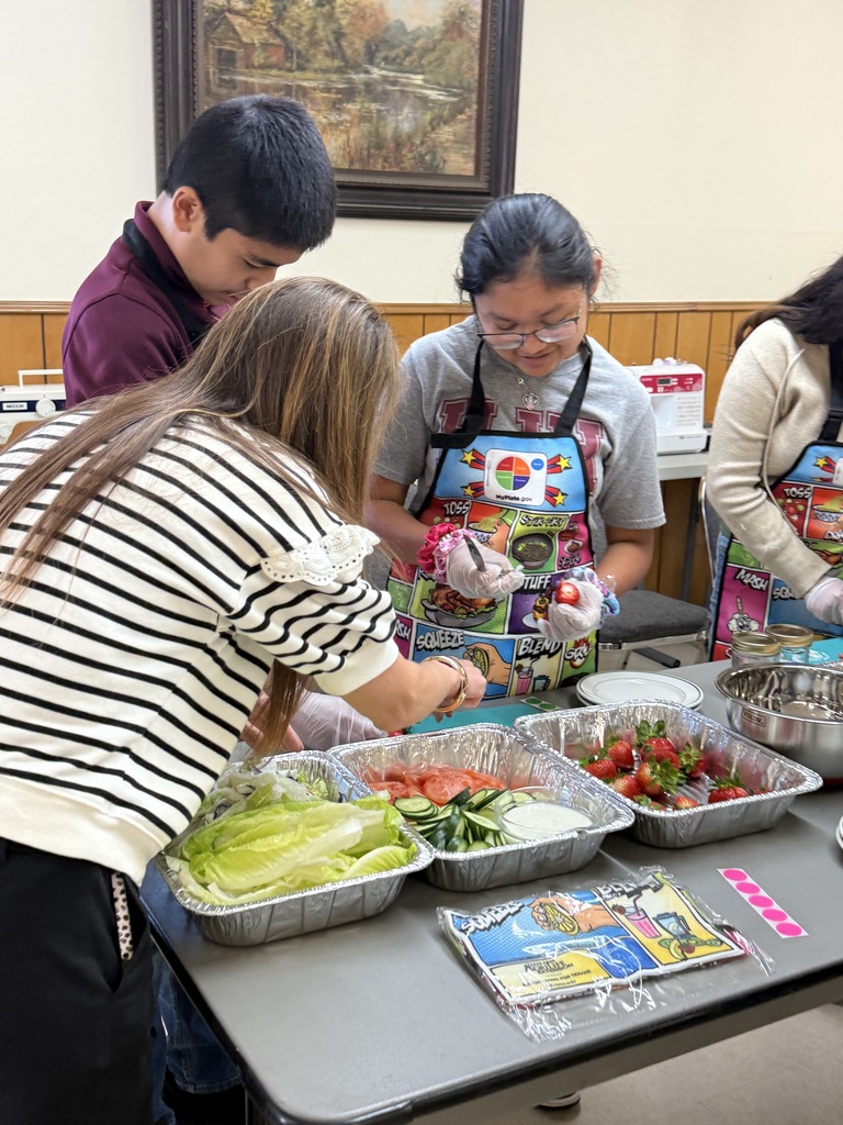 People wear aprons as they prepare food on a table. Bowls, plates, and a tray hold vegetables.