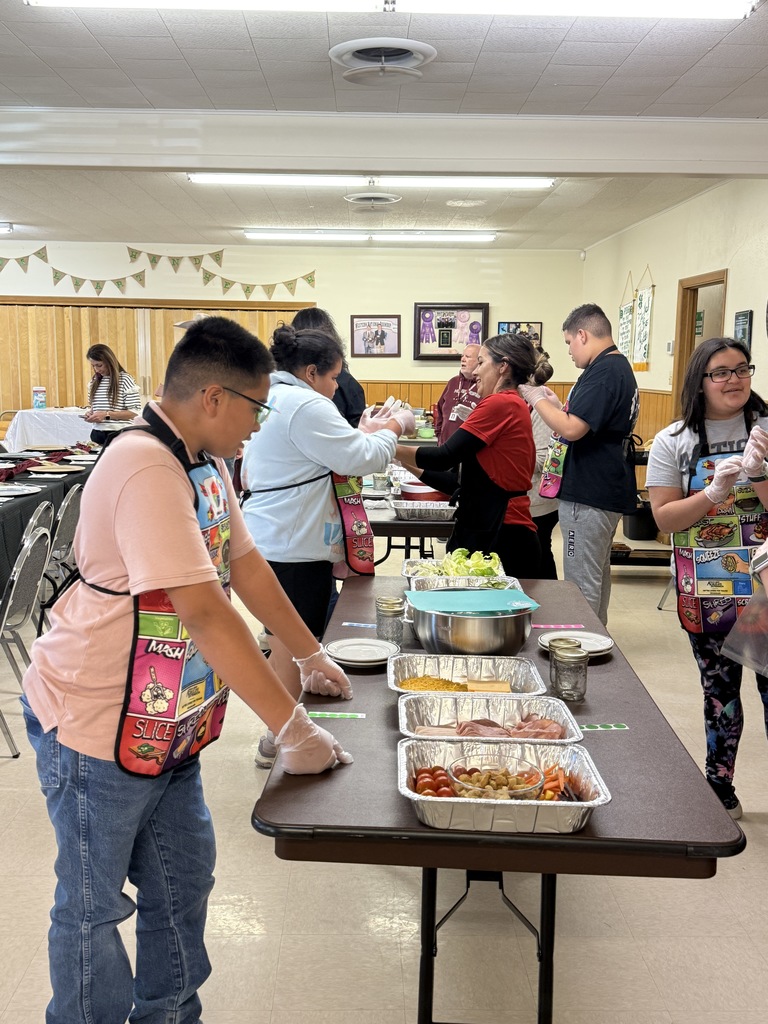 People in an indoor area with tables of food. One person stands with an apron, leaning over the table.