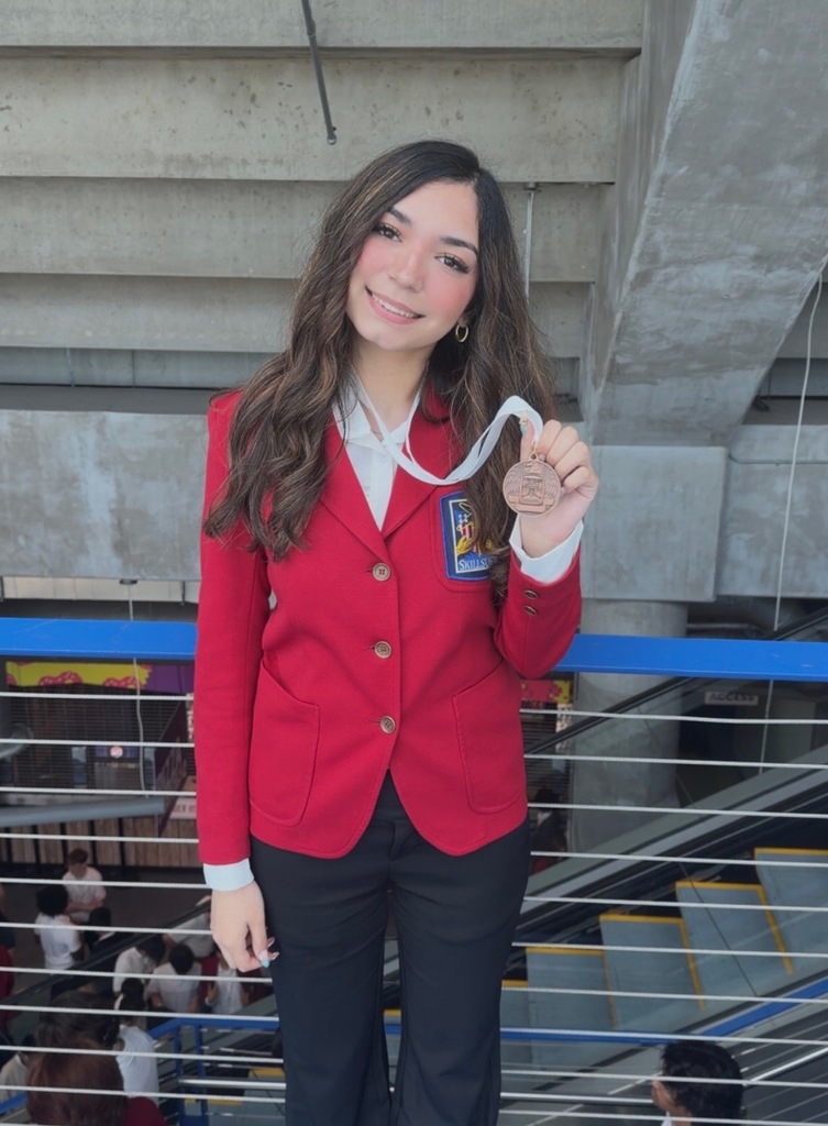 Woman in a red blazer with buttons and a medal in hand stands in front of stairs.