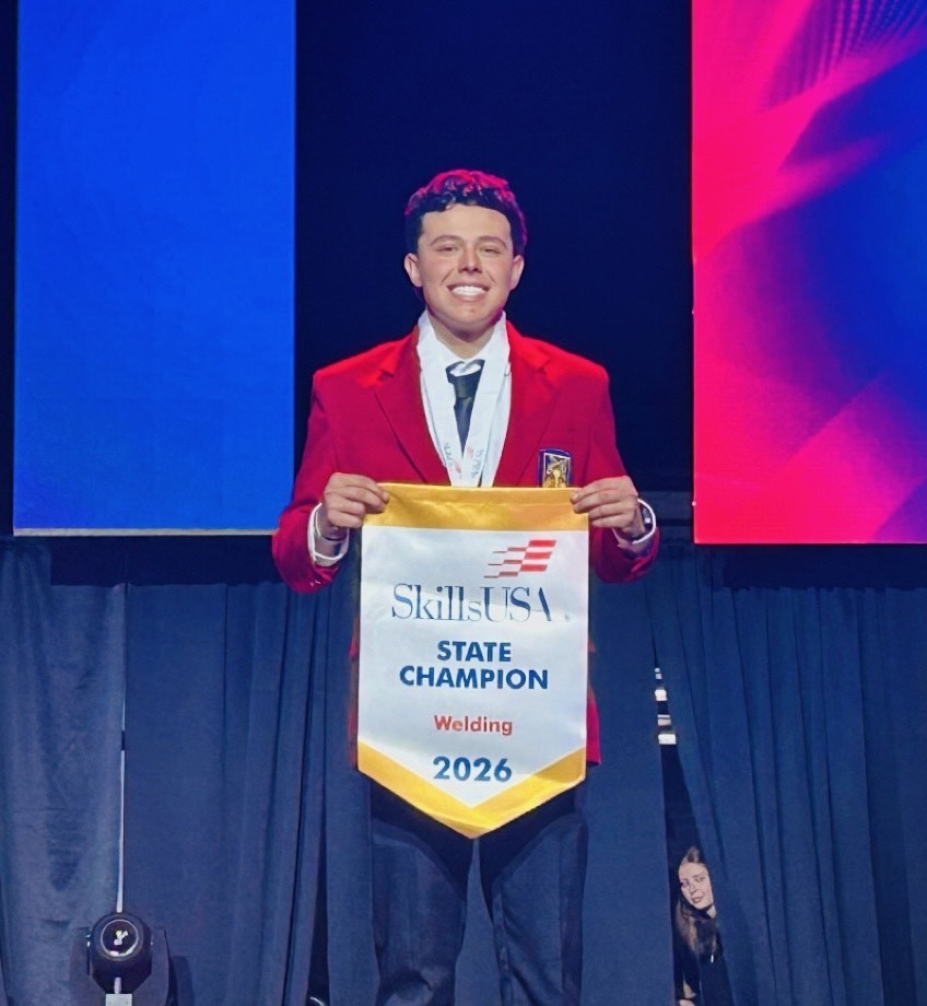Man wearing a red jacket and tie holds a banner that says State Champion Welding 2026.