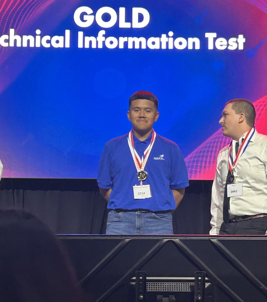 Two men wearing medals stand on a stage, with one looking at the camera. A large screen behind reads "GOLD Technical Information Test."