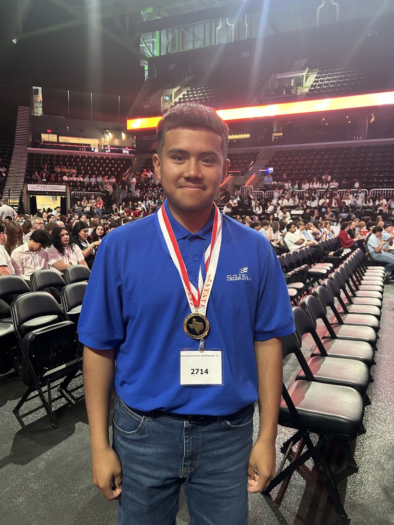 A young man stands in a stadium with a crowd, wearing a blue shirt, blue jeans, and a gold medal.