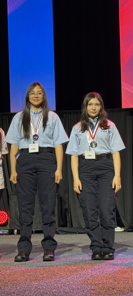 Two women, both in uniforms, stand side by side on stage, each wearing a medal around their necks.