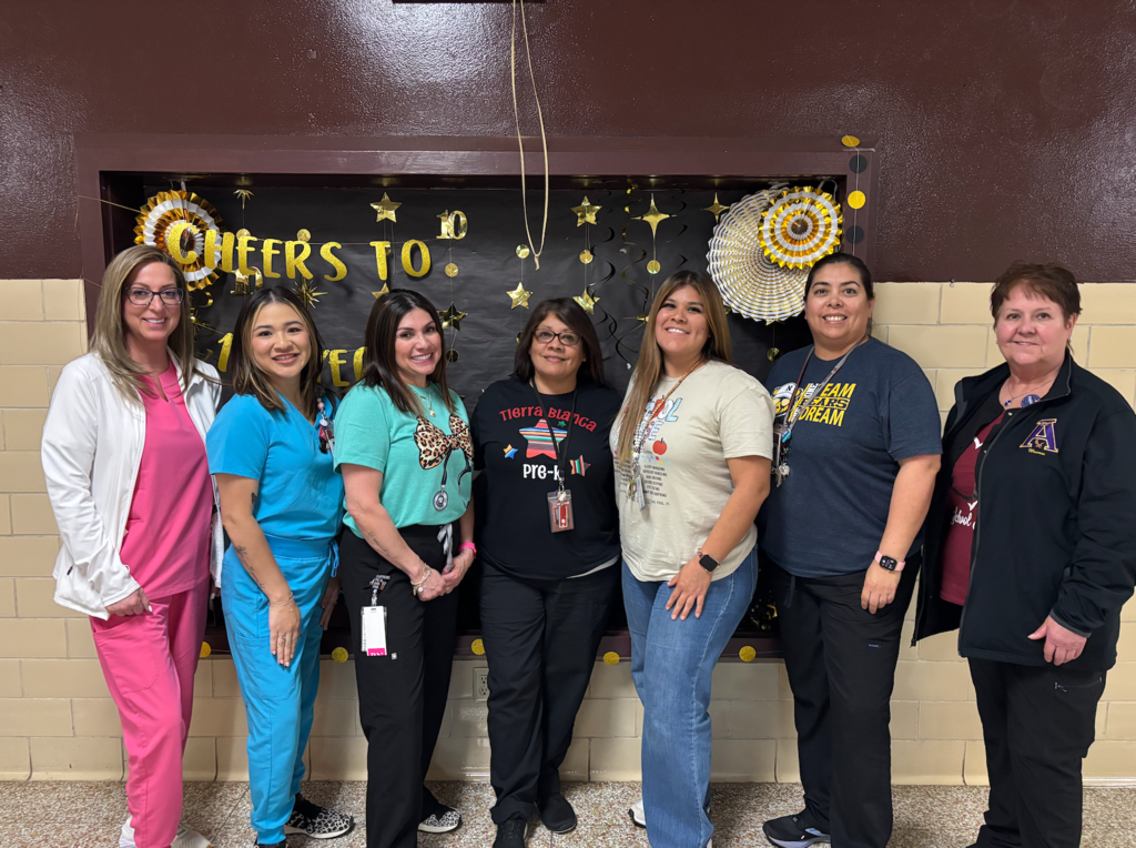 Six women stand close together in front of a decorative wall with a banner reading "CHEERS TO".