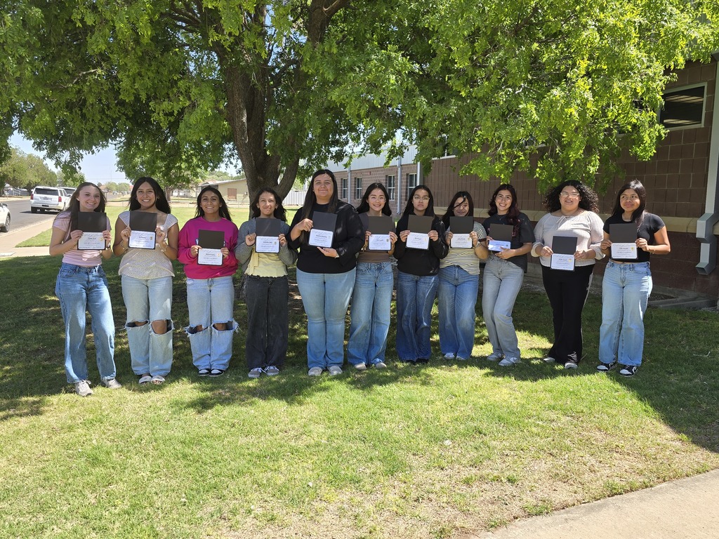 Group of students in matching shirts standing in front of a building, holding certificates, and posing for a photo.